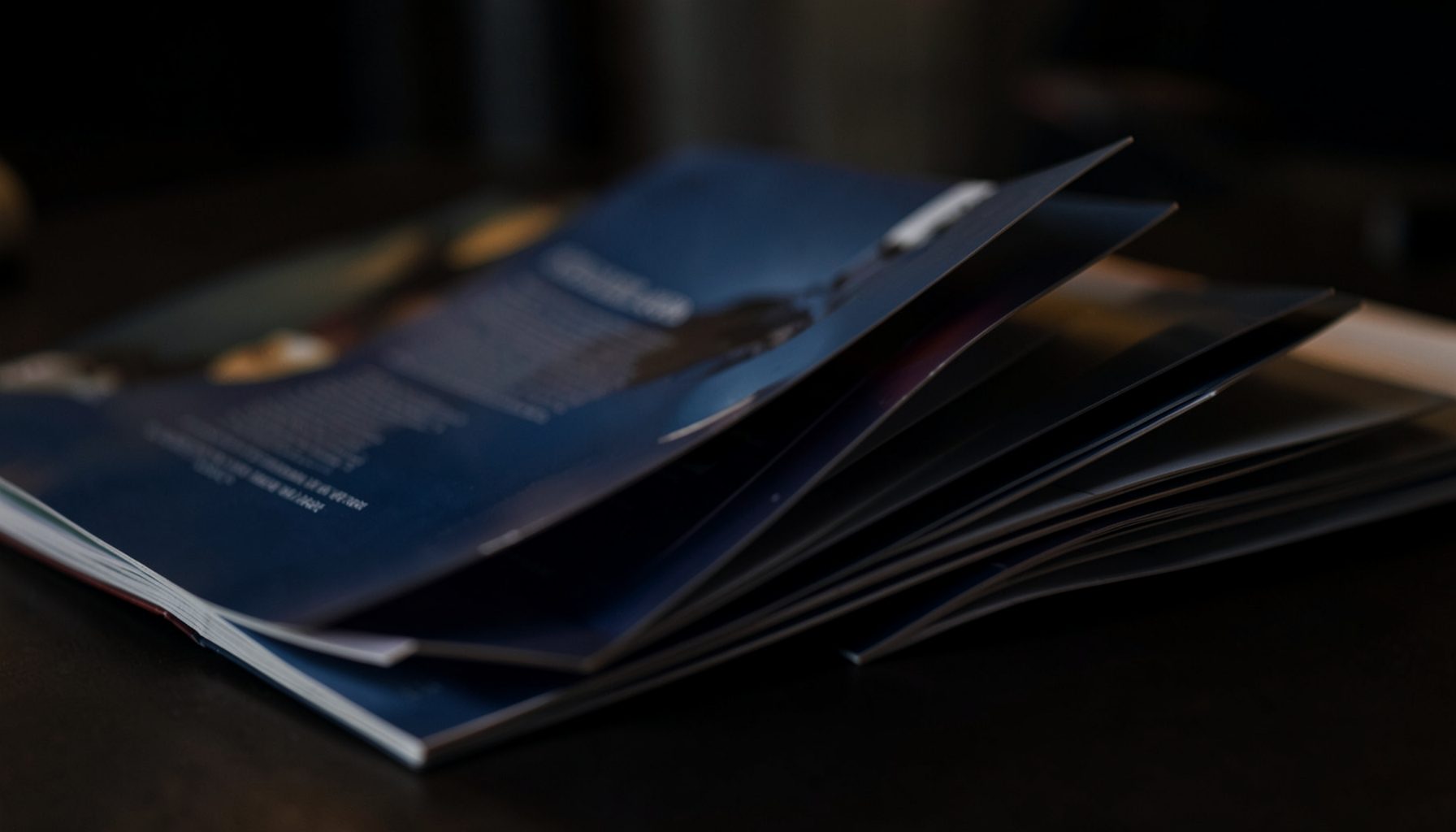 A dark, atmospheric close-up of glossy magazine pages fanned open on a desk, with deep navy and charcoal tones accented by soft warm highlights.